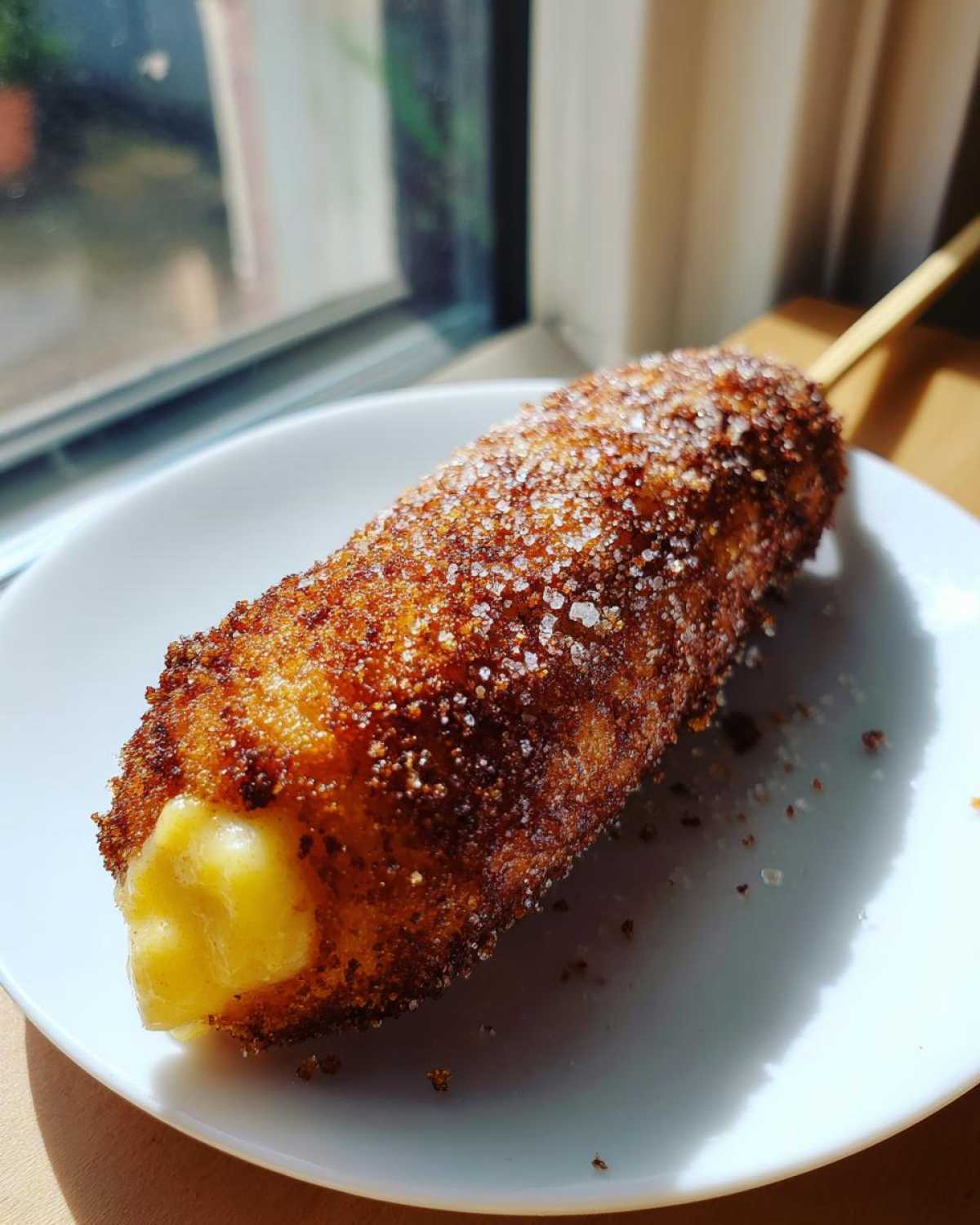 A crispy, sugar-dusted korean corn dog on a stick, showing melted cheese oozing from the end.