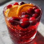 Close-up of a glass filled with sparkling red Thanksgiving punch, garnished with fresh cranberries and orange slices.