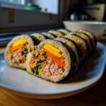 A row of freshly sliced tuna kimbap rolls showing layers of rice, tuna filling, carrots, and egg on a white plate.
