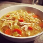 Close-up of a white bowl filled with steaming turkey noodle soup, featuring egg noodles, shredded turkey, carrots, and celery.
