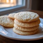A stack of four perfectly baked soft sugar cookies, lightly coated in sugar, sitting on a white plate.