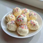 Eight white chocolate sugar cookie truffles topped with colorful sprinkles, arranged on a white plate.