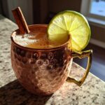 A fizzy apple cider moscow mule served in a hammered copper mug, garnished with a lime wheel and cinnamon stick.