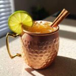 A frosty copper mug filled with an apple cider moscow mule, garnished with a lime wheel and cinnamon sticks.