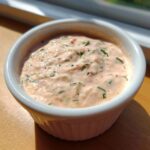 A close-up of creamy, pinkish remoulade sauce speckled with green herbs and small red pieces in a white ramekin.