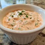 A close-up of a white ramekin filled with creamy, pinkish remoulade sauce, garnished with fresh parsley.