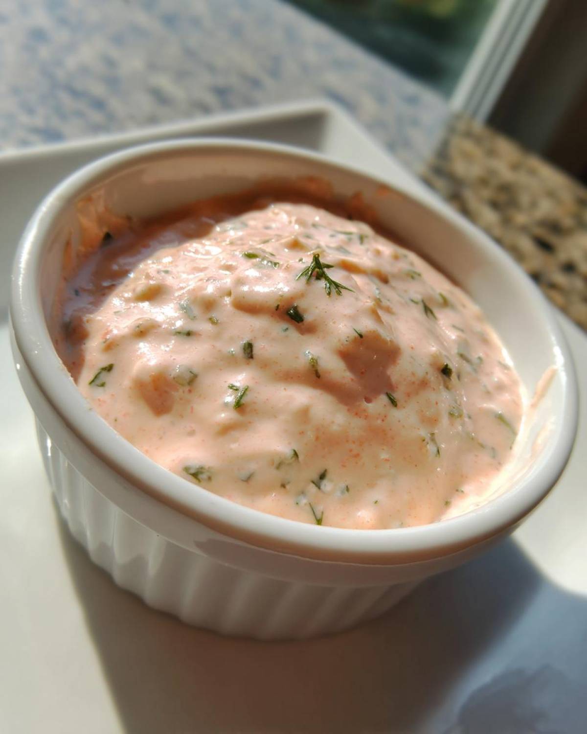 A close-up shot of creamy, pink remoulade sauce speckled with green herbs, served in a small white ramekin.