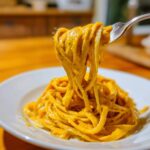 A fork lifting spaghetti coated thickly in bright orange butternut squash pasta sauce from a white plate.