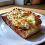A thick slice of cheesy egg toast, topped with melted and browned cheese and ricotta, sitting on a white plate.