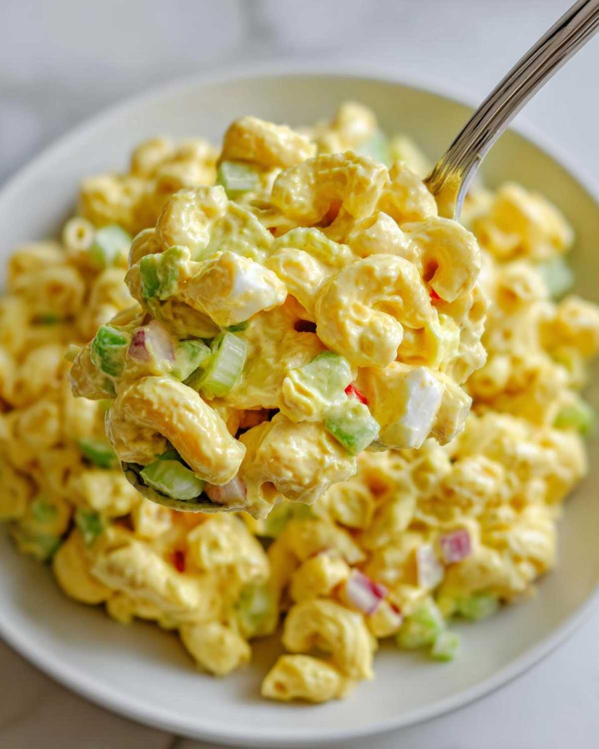 A fork lifting a generous scoop of creamy Copycat Deli-Style Macaroni Salad, showing elbow macaroni, celery, and egg pieces.