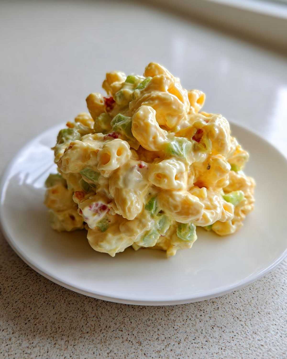 A mound of creamy Copycat Deli-Style Macaroni Salad featuring elbow macaroni, celery, and red pepper pieces on a white plate.