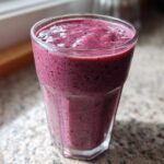 A close-up of a thick, vibrant purple Creamy Blueberry Banana Smoothie served in a clear glass on a speckled countertop.
