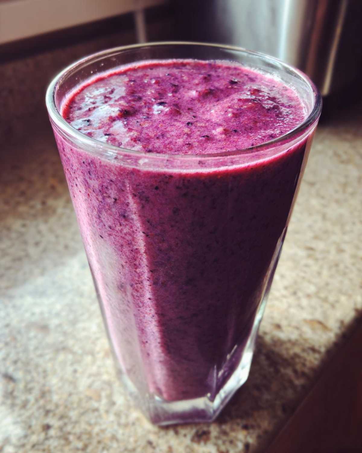 A close-up of a tall glass filled with a thick, vibrant purple Creamy Blueberry Banana Smoothie resting on a speckled countertop.