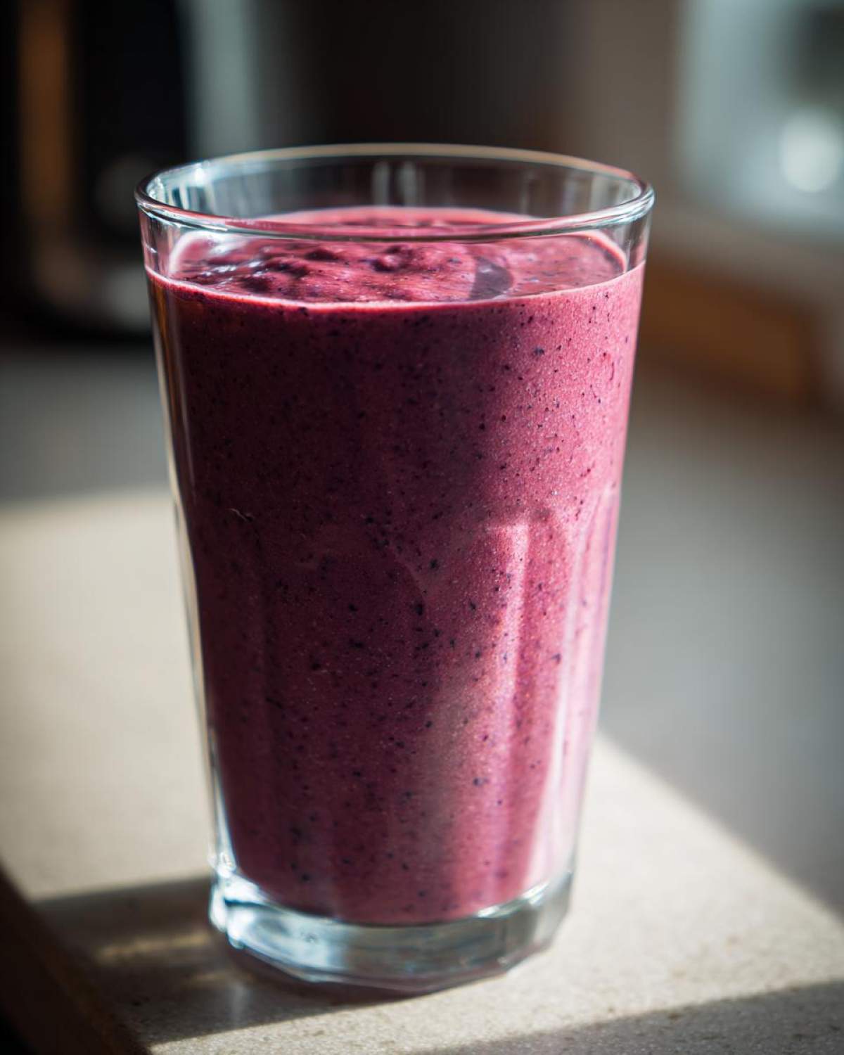 A tall glass filled with a thick, deep purple Creamy Blueberry Banana Smoothie, sitting on a countertop in natural light.