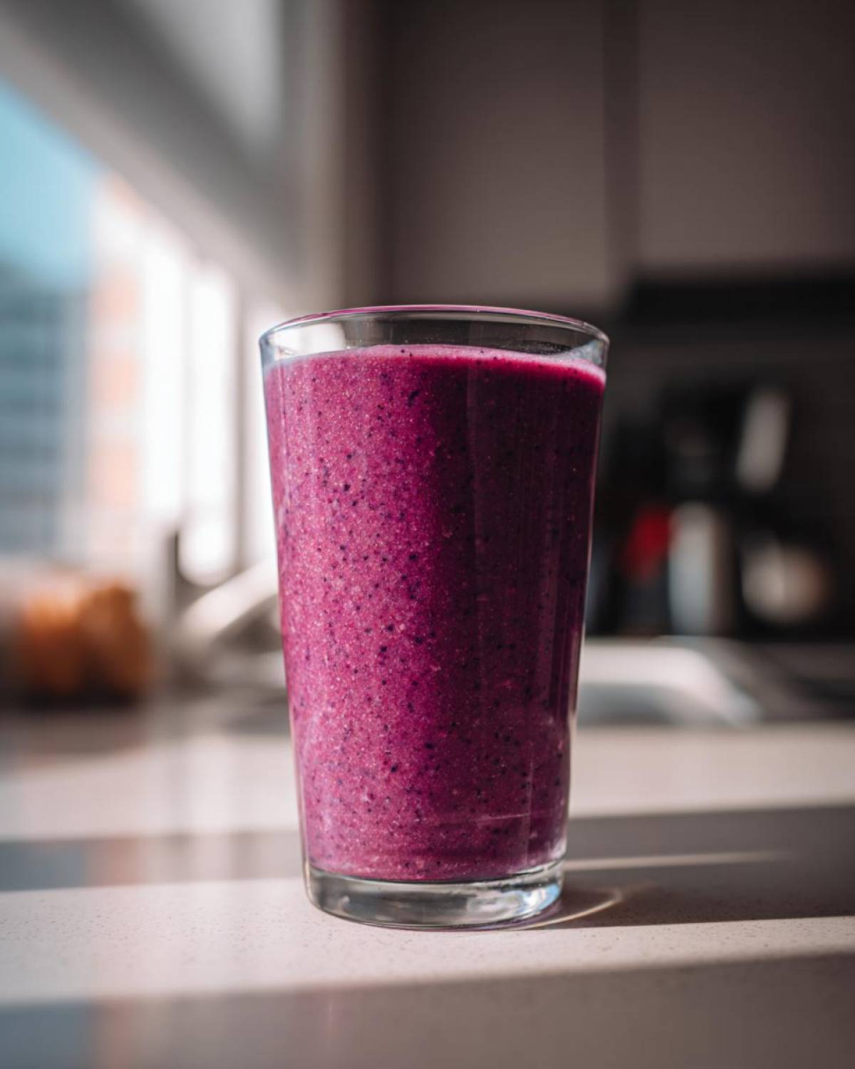 A tall glass filled with a thick, vibrant purple Creamy Blueberry Banana Smoothie sitting on a kitchen counter.