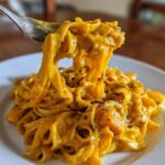 A fork lifts creamy fettuccine coated in vibrant butternut squash pasta sauce from a white plate.