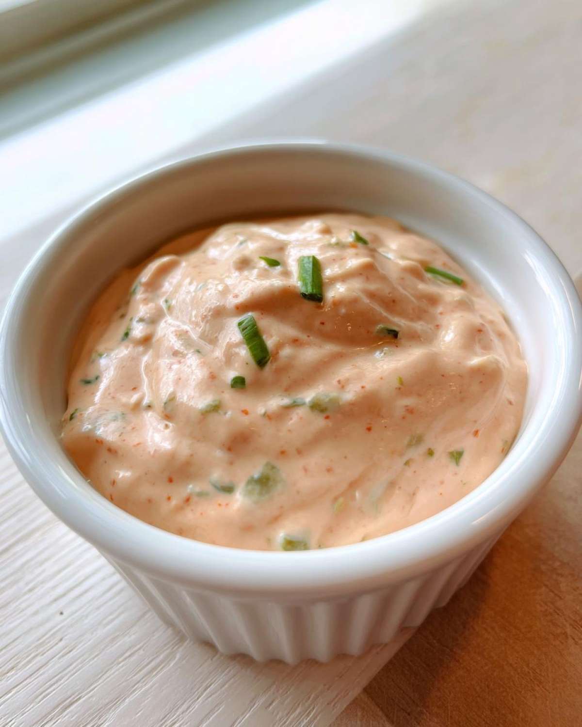 Close-up of a small white bowl filled with thick, pinkish-orange remoulade sauce, garnished with chopped green chives.