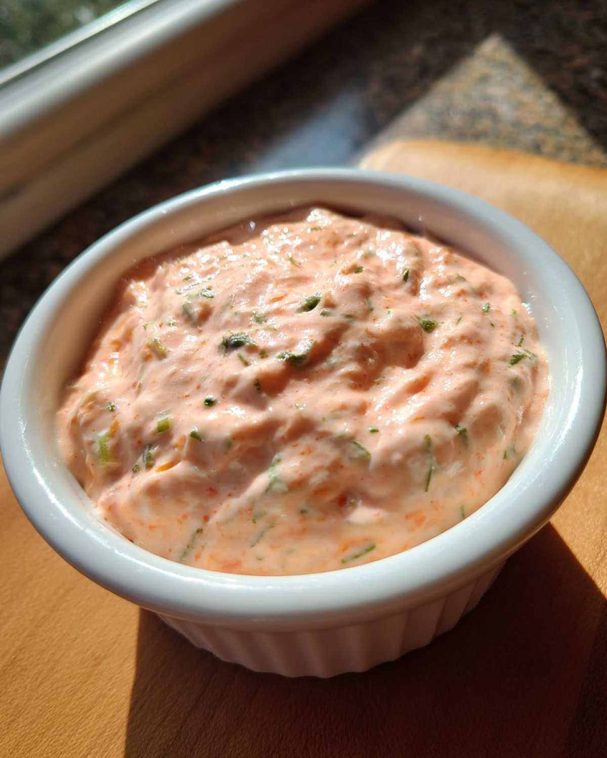 A close-up of creamy, orange-tinted remoulade sauce speckled with green herbs, served in a white dipping bowl.