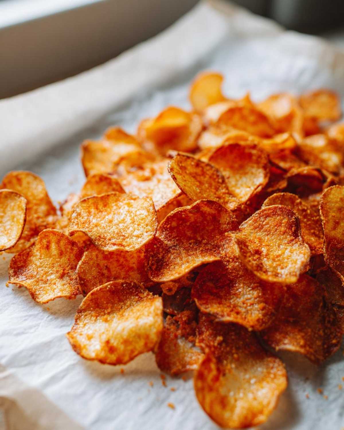 Close-up of a pile of golden brown, crispy oven Parmesan potato chips scattered on white parchment paper.