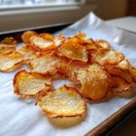 A close-up of golden, crispy oven Parmesan potato chips piled on parchment paper on a baking sheet.