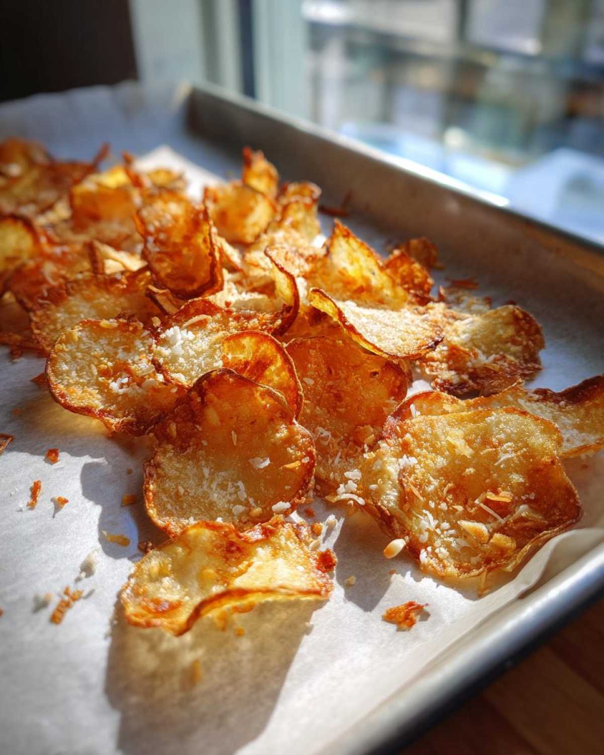 Golden brown, crispy oven Parmesan potato chips scattered on parchment paper on a baking sheet, catching bright sunlight.