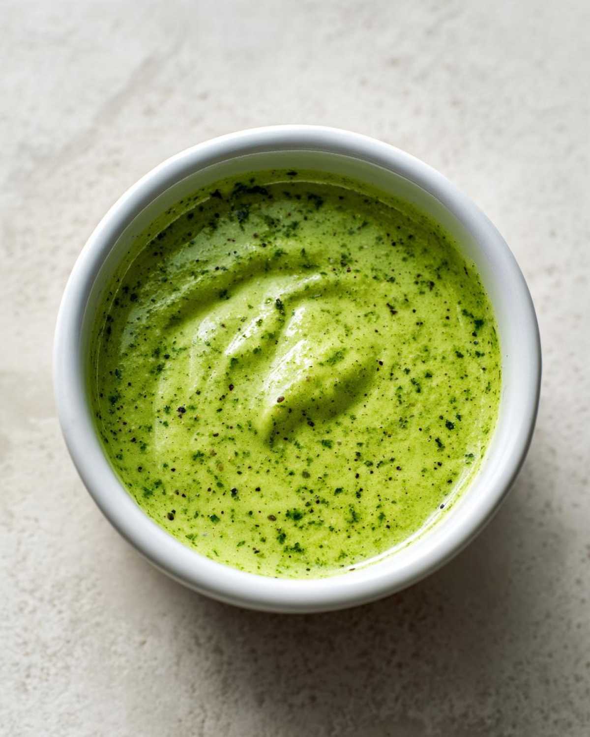 Overhead shot of creamy, bright green Five-Minute Avocado Lime Dressing speckled with herbs in a small white ramekin.