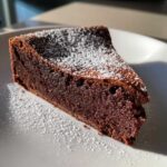 A close-up slice of dense, dark flourless chocolate torte dusted generously with white powdered sugar on a white plate.
