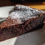 A close-up of a dark, dense slice of flourless chocolate torte dusted with white powdered sugar on a white plate.