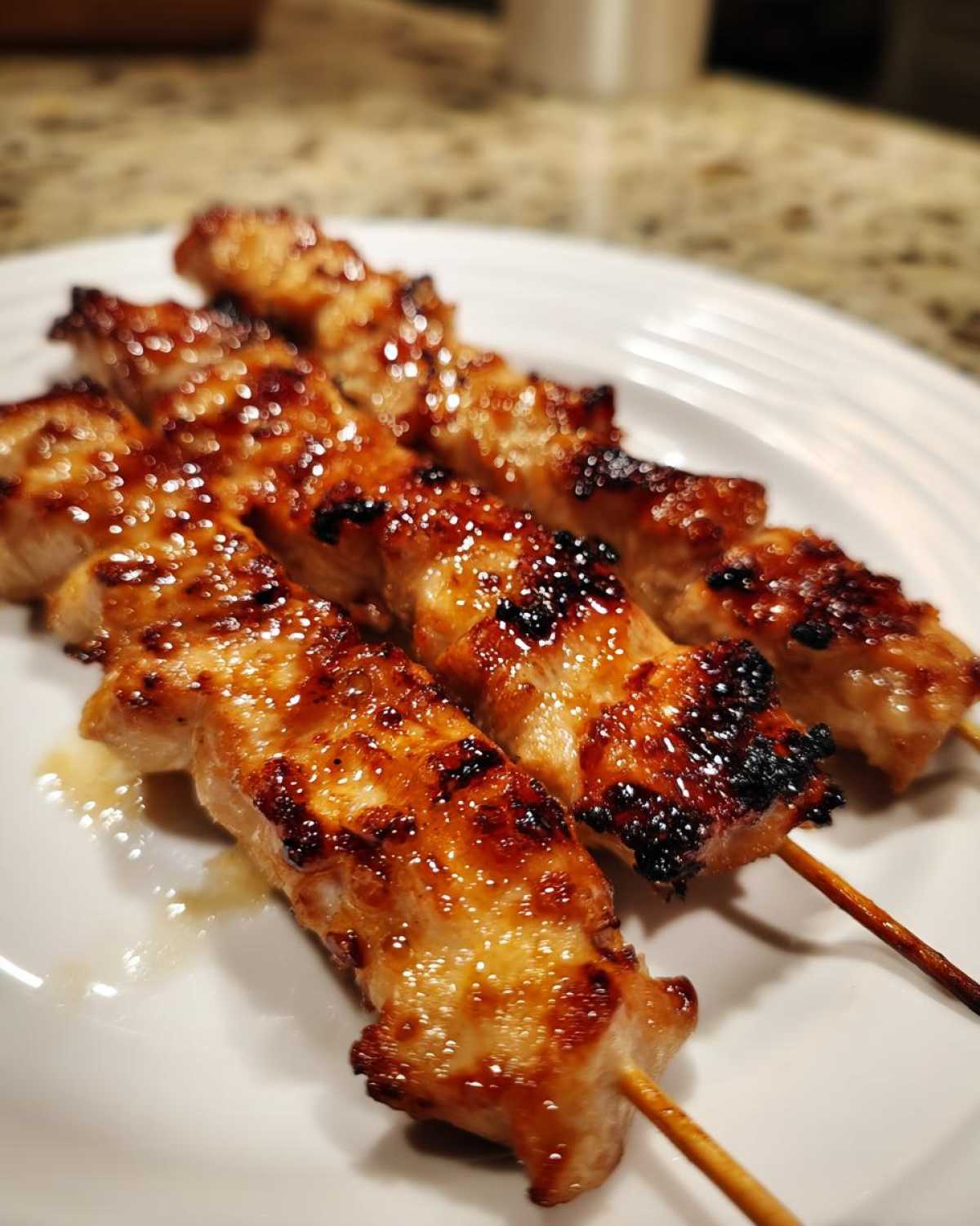 Close-up of three glazed and slightly charred chicken skewers resting on a white plate.