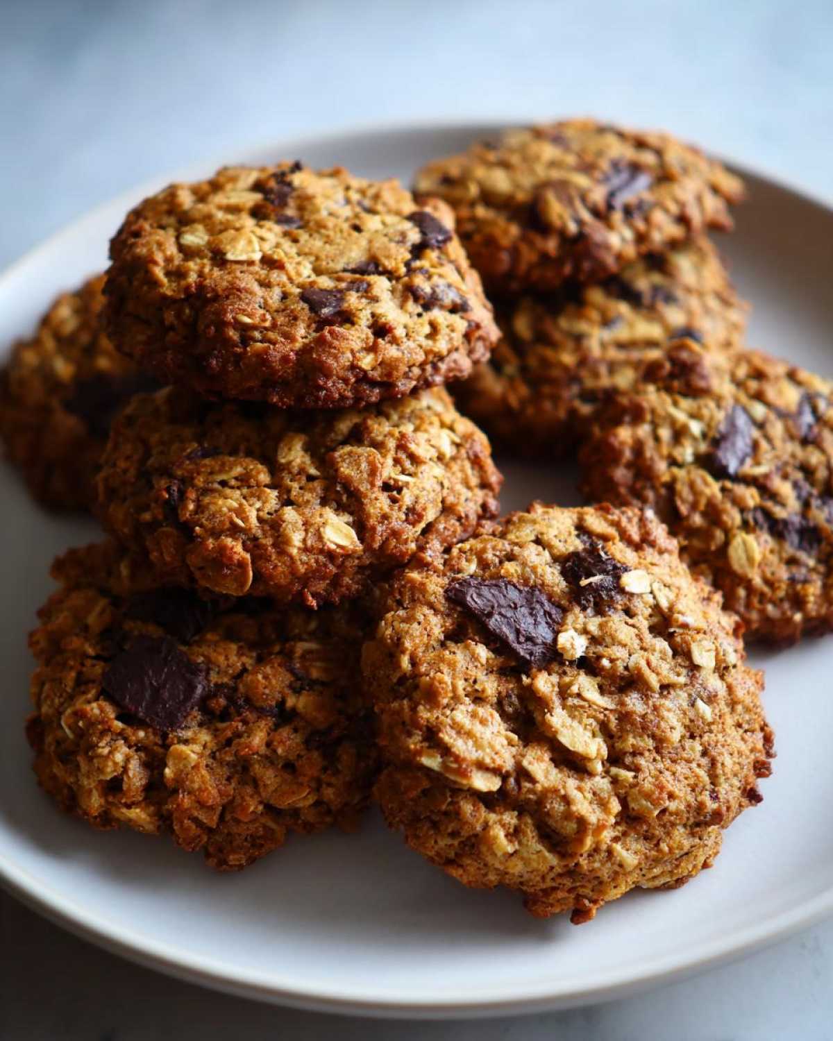 A close-up of several chewy Gluten-Free Irish Oat Cookies with dark chocolate chunks stacked on a light plate.