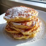 A tall stack of fluffy High-Protein Cottage Cheese Pancakes dusted generously with powdered sugar on a white plate.