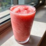 A tall glass filled with a bright pink, icy watermelon smoothie sitting on a sunlit windowsill.