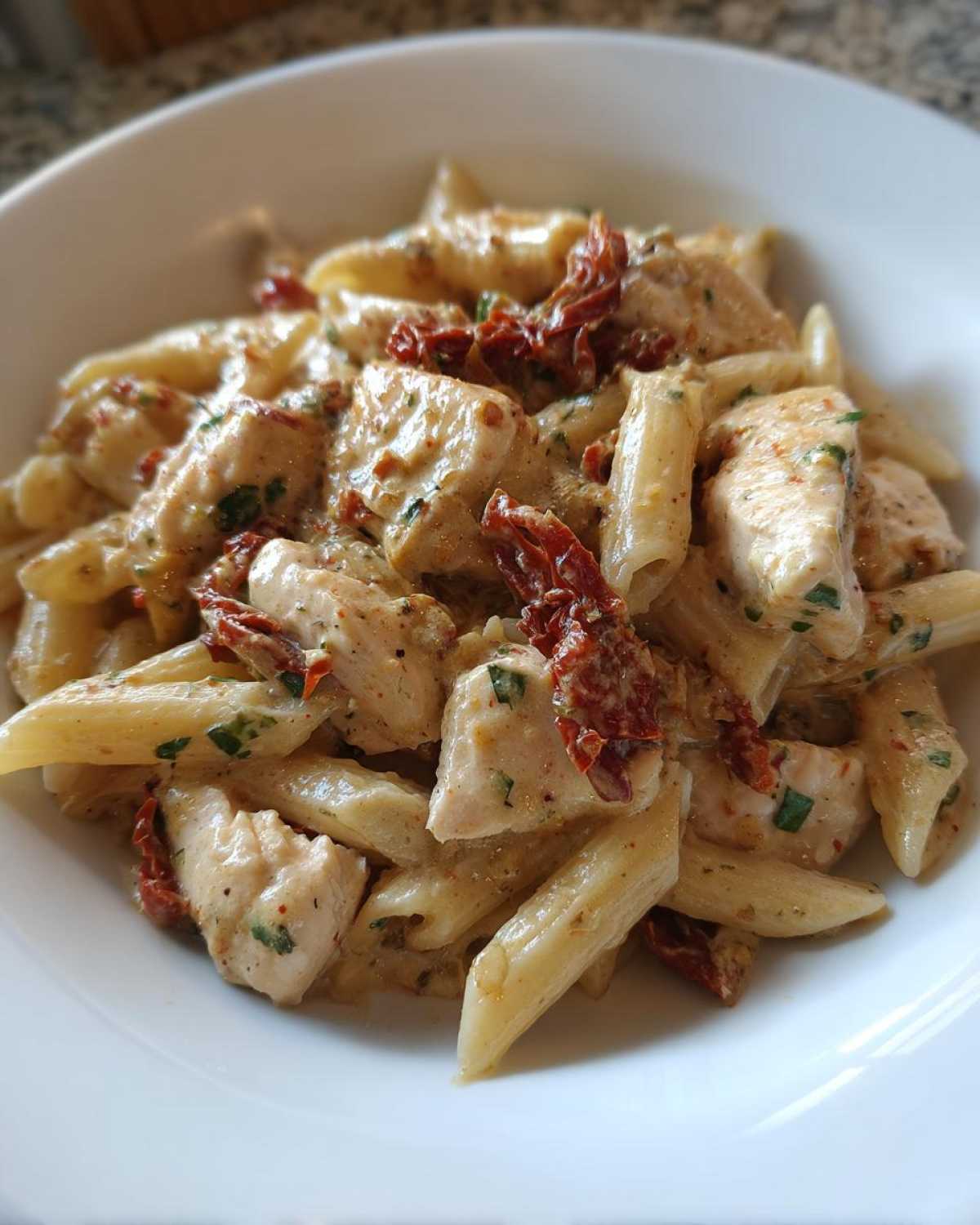 A close-up serving of creamy marry me chicken pasta with penne, chicken chunks, and sun-dried tomatoes in a white bowl.