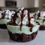 A close-up of a No-Bake Mint Chocolate Cheesecake Cup featuring a dark crust, light green filling, whipped cream, and chocolate drizzle.