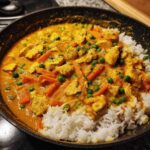 Close-up of One-Pot Coconut Curry Chicken and Rice with carrots and peas served over white rice in a black skillet.