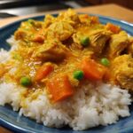 A close-up of a blue bowl filled with fluffy white rice topped with rich, yellow One-Pot Coconut Curry Chicken and Rice, featuring carrots and peas.