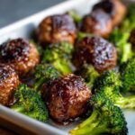Close-up of glazed Sheet Pan Ginger Sesame Chicken Meatballs with roasted broccoli florets on a baking sheet.