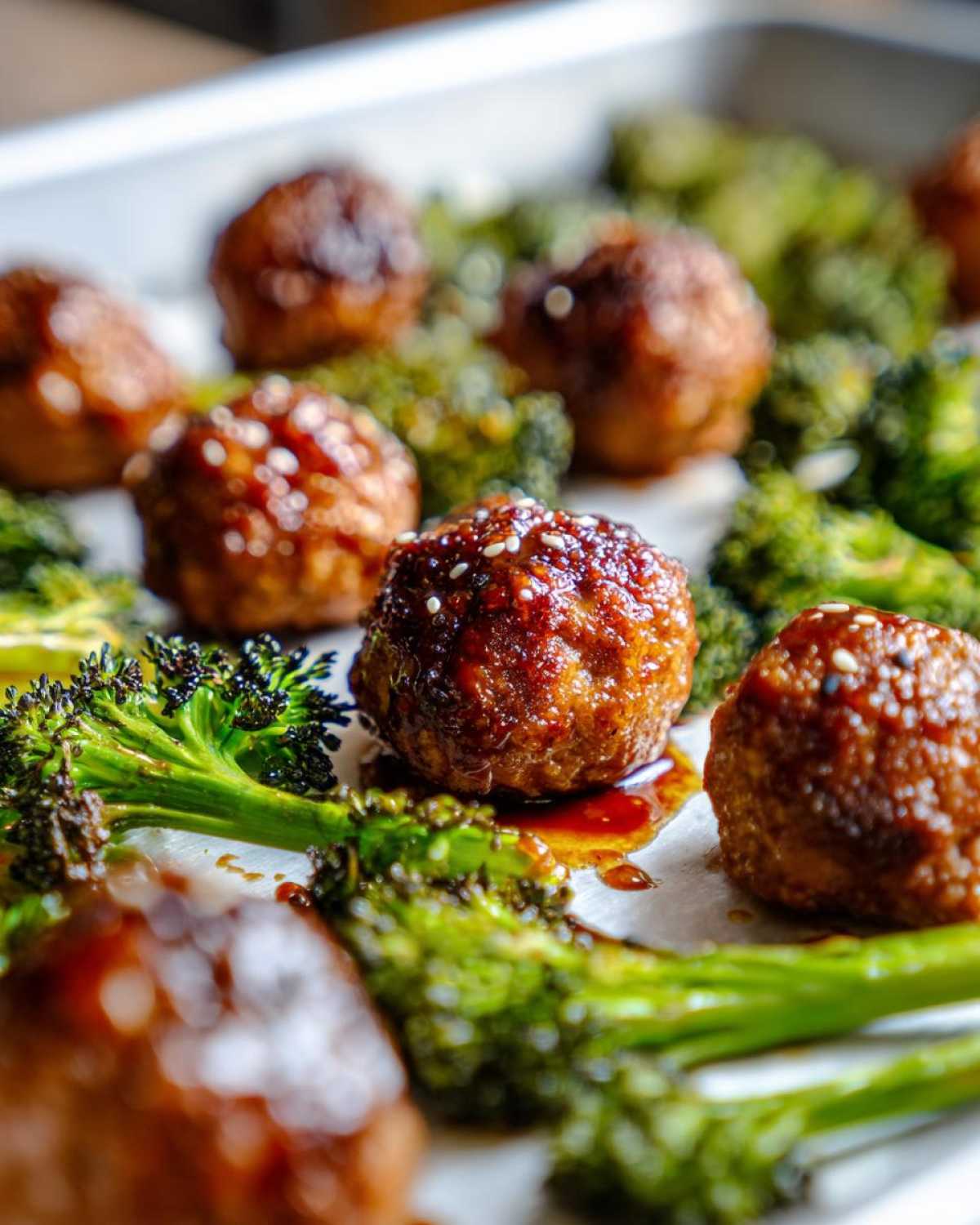 Close-up of glazed Sheet Pan Ginger Sesame Chicken Meatballs with roasted broccoli florets on a baking sheet.