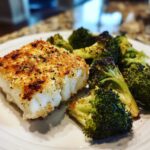 A flaky piece of Sheet Pan Parmesan Crusted Cod served next to roasted broccoli florets on a white plate.