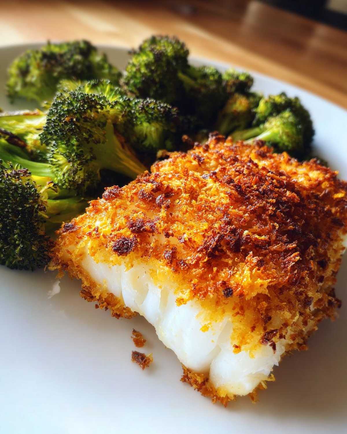 Close-up of a flaky white cod fillet with a golden, crispy Parmesan crust next to roasted broccoli florets.