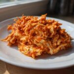 Close-up of a mound of shredded buffalo chicken coated in vibrant orange sauce on a white plate.