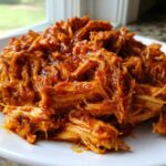 Close-up of saucy, shredded buffalo chicken piled high on a white plate.
