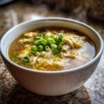 A close-up of silky egg drop soup with wisps of cooked egg in savory broth, topped with fresh chopped green onions.