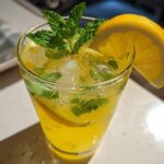 Close-up of a tall glass filled with a yellow Sparkling Lemon Mint Mocktail, ice, mint leaves, and a lemon slice garnish.
