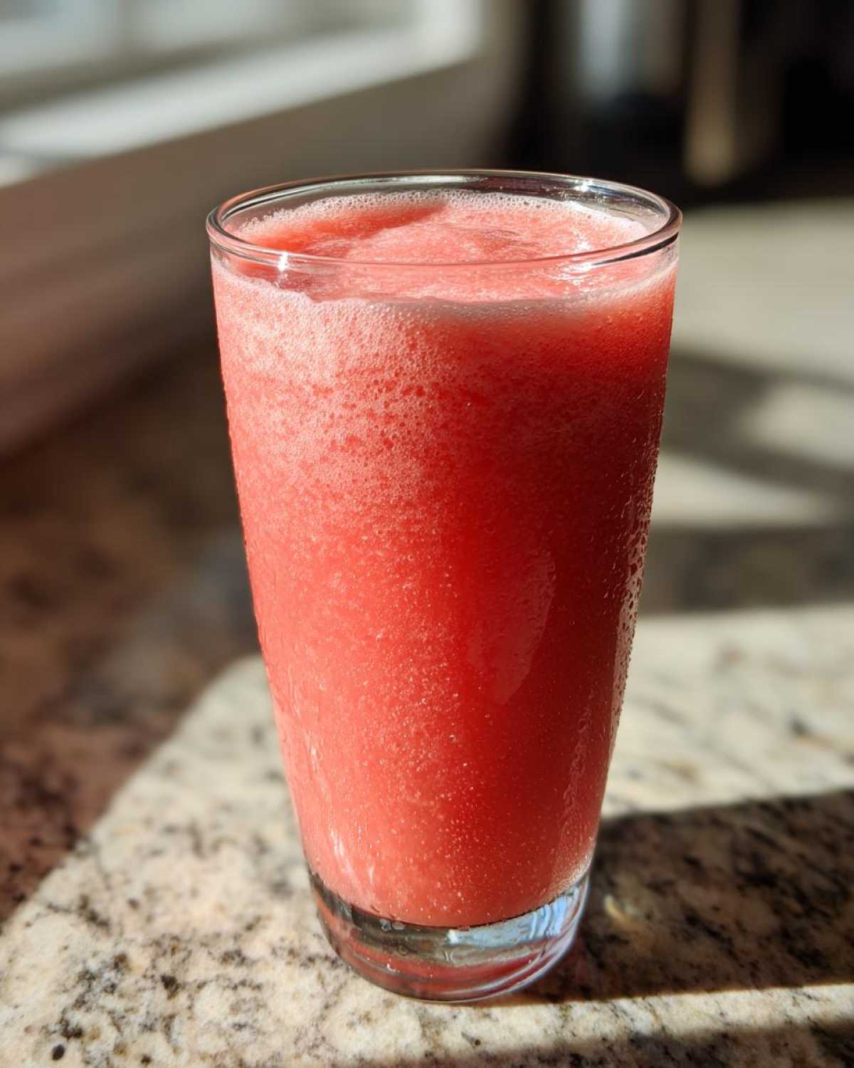 A tall glass filled with a vibrant, frothy watermelon smoothie sitting on a granite countertop in bright sunlight.