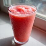 A tall glass filled with a bright red, icy watermelon smoothie sitting on a sunlit windowsill.