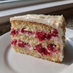 A close-up slice of white chocolate raspberry cake showing two layers filled generously with raspberries and white frosting.