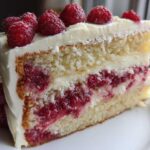 A close-up of a moist slice of white chocolate raspberry cake layered with white frosting and fresh raspberries.