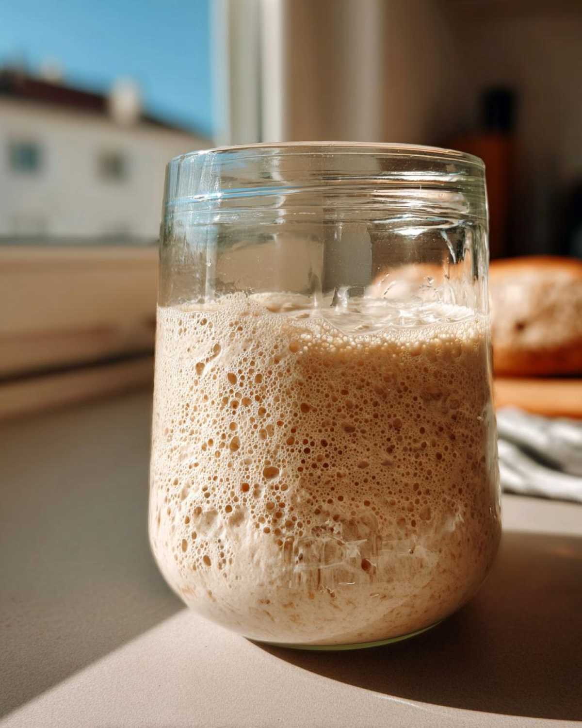 A highly active sourdough starter, full of bubbles, resting in a clear glass jar near a sunny window.