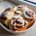 A close-up of freshly baked cinnamon buns covered in thick white icing, sitting in a white dish.